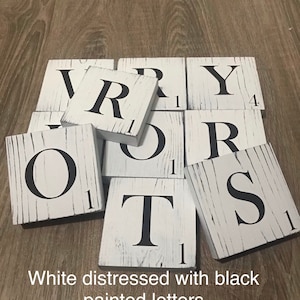 Op de afbeelding: Witte, verweerde houten tegels met zwart geschilderde letters die het woord "VERY" spellen. Elke tegel heeft een Scrabble-achtig nummer in de hoek. De tekst "White distressed with black painted letters" staat onderaan.