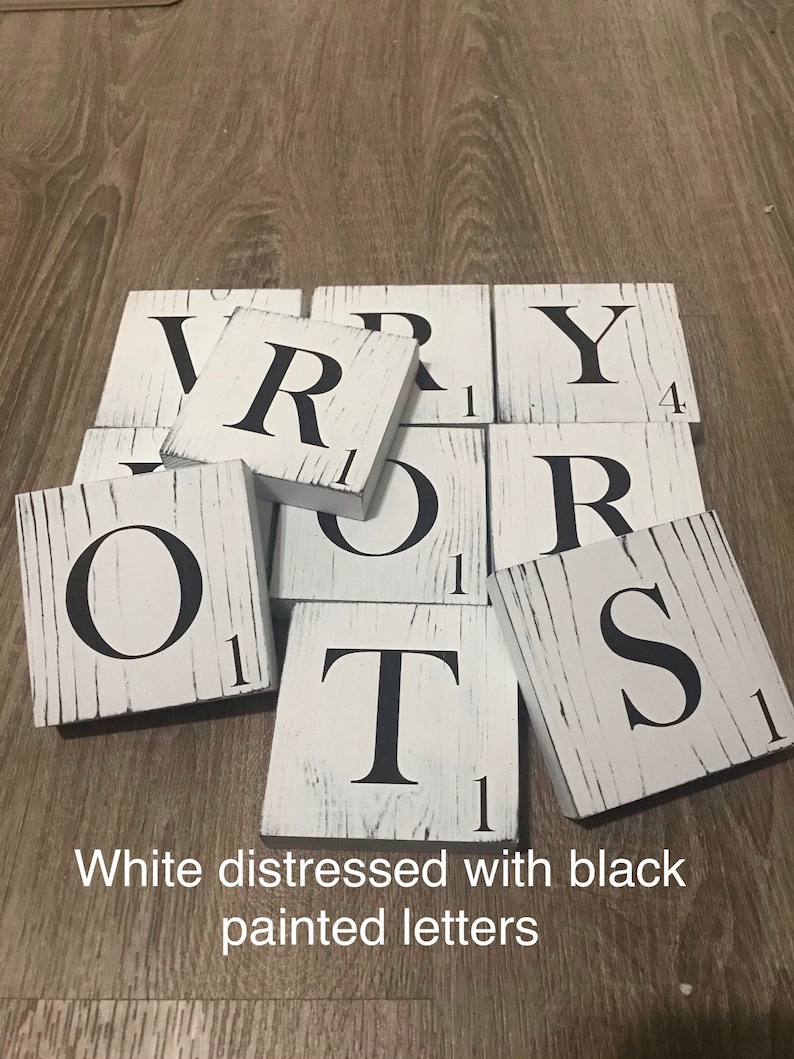 Op de afbeelding: Witte, verweerde houten tegels met zwart geschilderde letters die het woord "VERY" spellen. Elke tegel heeft een Scrabble-achtig nummer in de hoek. De tekst "White distressed with black painted letters" staat onderaan.