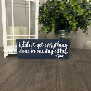 May include: A dark blue wooden sign with white lettering that reads "I didn't get everything done in one day either. -God".