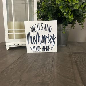 May include: A square white wooden sign with dark blue text that reads MEALS AND memories MADE HERE, featuring a small heart symbol on each side of the bottom line of text. The sign is positioned upright on a dark brown wooden surface.