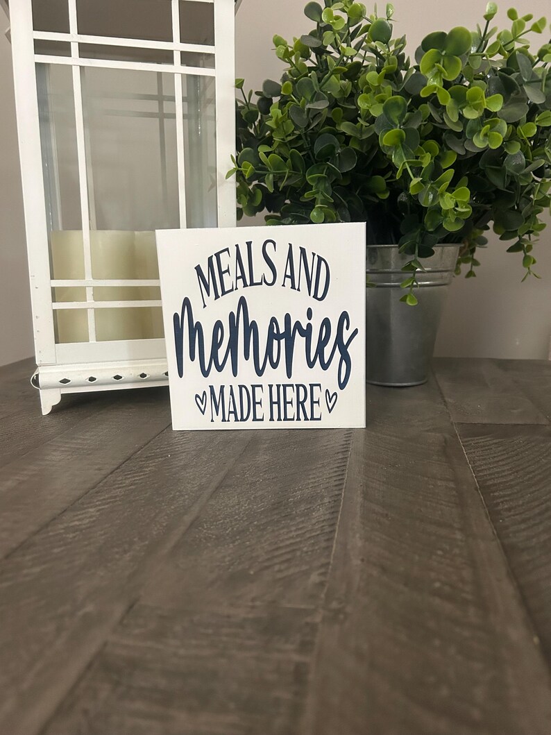 May include: A square white wooden sign with dark blue text that reads MEALS AND memories MADE HERE, featuring a small heart symbol on each side of the bottom line of text. The sign is positioned upright on a dark brown wooden surface.