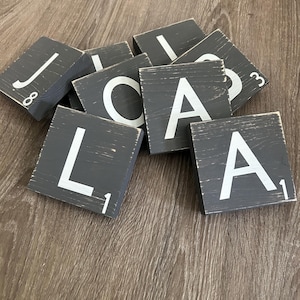 May include: A collection of gray wooden letter tiles with white letters and numbers. The letters include J, L, A, and C. The tiles are arranged on a wooden surface.