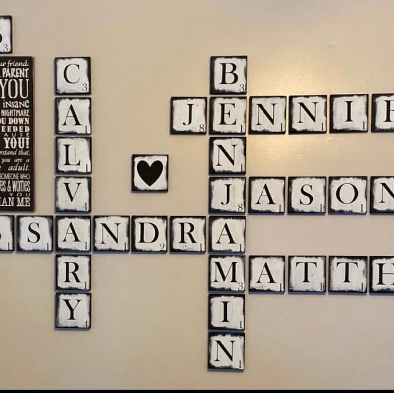 Op de afbeelding: Een wanddecoratie van individuele houten tegels, die lijken op Scrabble-stukken. De tegels spellen namen en zinnen, met een zwarte harttegel. De tegels zijn wit met zwarte letters en randen.