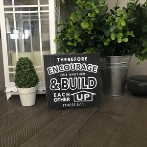 May include: Black wooden sign with white text that reads "Therefore Encourage One Another & Build Each Other Up 1 Thess 5:11".