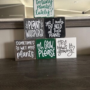 May include: A collection of five square wooden signs with distressed edges. The top sign is white with green text reading Botany plants lately?. The middle signs are black with white text reading Plant Whisperer and yes I really DO NEED all these plants. The bottom signs are brown, green, and white with text reading SOMETIMES I WET MY plants, OH GROW... PLEASE, and HEY How's It growing?
