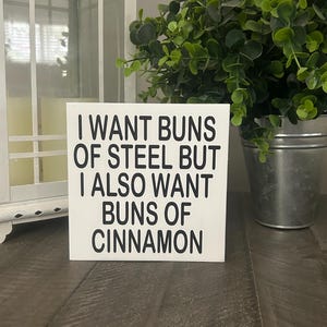 May include: A square white wooden sign with black text that reads I WANT BUNS OF STEEL BUT I ALSO WANT BUNS OF CINNAMON. The sign features a clean, flat surface with bold, sans-serif lettering centered on the front, presenting a humorous message.