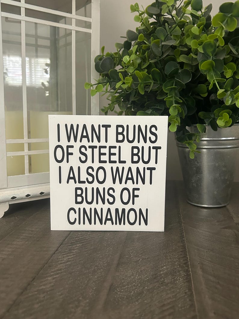 May include: A square white wooden sign with black text that reads I WANT BUNS OF STEEL BUT I ALSO WANT BUNS OF CINNAMON. The sign features a clean, flat surface with bold, sans-serif lettering centered on the front, presenting a humorous message.