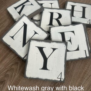 Op de afbeelding: Een verzameling witgrijze houten lettertegels met zwarte hoofdletters. De tegels hebben een verweerde uitstraling en zijn gerangschikt op een houten oppervlak. Sommige tegels hebben nummers in de hoek. De tekst "Whitewash gray with black font" staat onderaan.