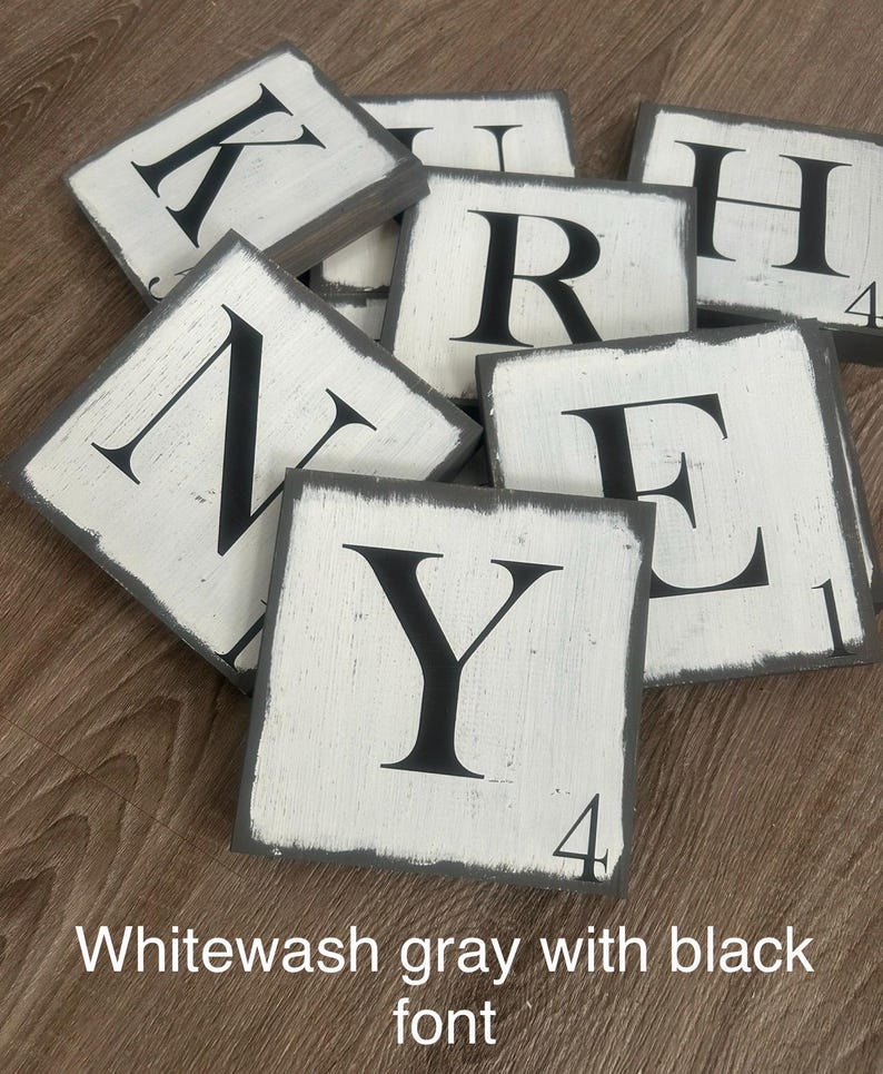Op de afbeelding: Een verzameling witgrijze houten lettertegels met zwarte hoofdletters. De tegels hebben een verweerde uitstraling en zijn gerangschikt op een houten oppervlak. Sommige tegels hebben nummers in de hoek. De tekst "Whitewash gray with black font" staat onderaan.