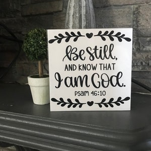 May include: White wooden sign with black lettering that reads "Be still, and know that I am God. Psalm 46:10". The sign is decorated with black floral designs and hearts.