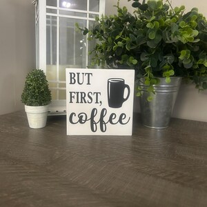 May include: A square white wooden sign featuring the black text "BUT FIRST, coffee" alongside a black graphic of a coffee mug. The sign is positioned in the center, displaying clear typography and a simple, bold design for home decor display.