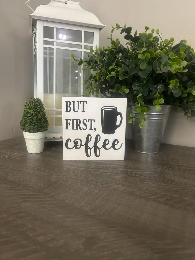 May include: A square white wooden sign featuring the black text "BUT FIRST, coffee" alongside a black graphic of a coffee mug. The sign is positioned in the center, displaying clear typography and a simple, bold design for home decor display.