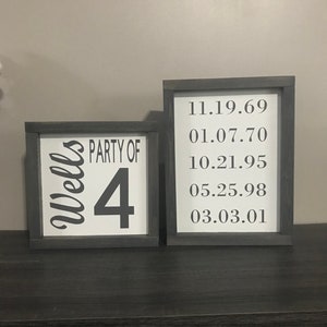 May include: Two framed signs with black frames. The sign on the left says "Wells Party of 4". The sign on the right has a list of dates: 11.19.69, 01.07.70, 10.21.95, 05.25.98, 03.03.01.