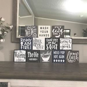 Puede incluir: Un conjunto de 12 letreros de baño de madera con varios refranes humorísticos. Los letreros están pintados de negro con letras blancas. Algunos de los refranes incluyen "Ready Aim Fire!" "The Ole Bath House" "Put a Lid On It" y "It Won't Flush Itself".