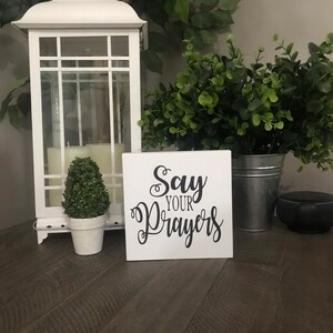 May include: White wooden sign with black calligraphy that reads "Say Your Prayers". The sign is sitting on a wooden table with a small potted plant and a white lantern in the background.