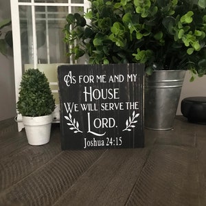May include: A square black wooden sign with distressed edges featuring white text that reads: As for me and my house we will serve the Lord. Joshua 24:15. The sign includes white leaf illustrations on both sides of the word Lord.