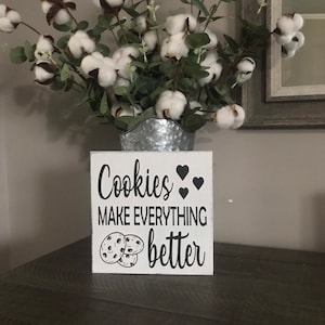 Może przedstawiać: Biały drewniany znak z czarnym tekstem, który brzmi "Cookies make everything better" z grafiką trzech ciasteczek z kawałkami czekolady. Znak stoi na ciemnobrązowej drewnianej powierzchni z wazą z bawełnianymi kwiatami i zielenią z tyłu.