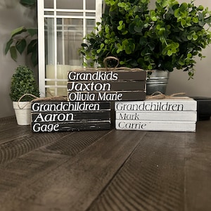 May include: Three wooden blocks stacked on top of each other. The top block is brown and says "Grandkids" in white lettering. The middle block is brown and says "Jaxton" in white lettering. The bottom block is brown and says "Olivia Marie" in white lettering. The second set of blocks is black and says "Grandchildren" in white lettering. The top block says "Aaron" in white lettering. The bottom block says "Gage" in white lettering. The third set of blocks is white and says "Grandchildren" in black lettering. The top block says "Mark" in black lettering. The bottom block says "Carrie" in black lettering.