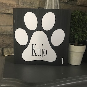 May include: Black wooden sign with a white paw print and the name "Kujo" written in white lettering.