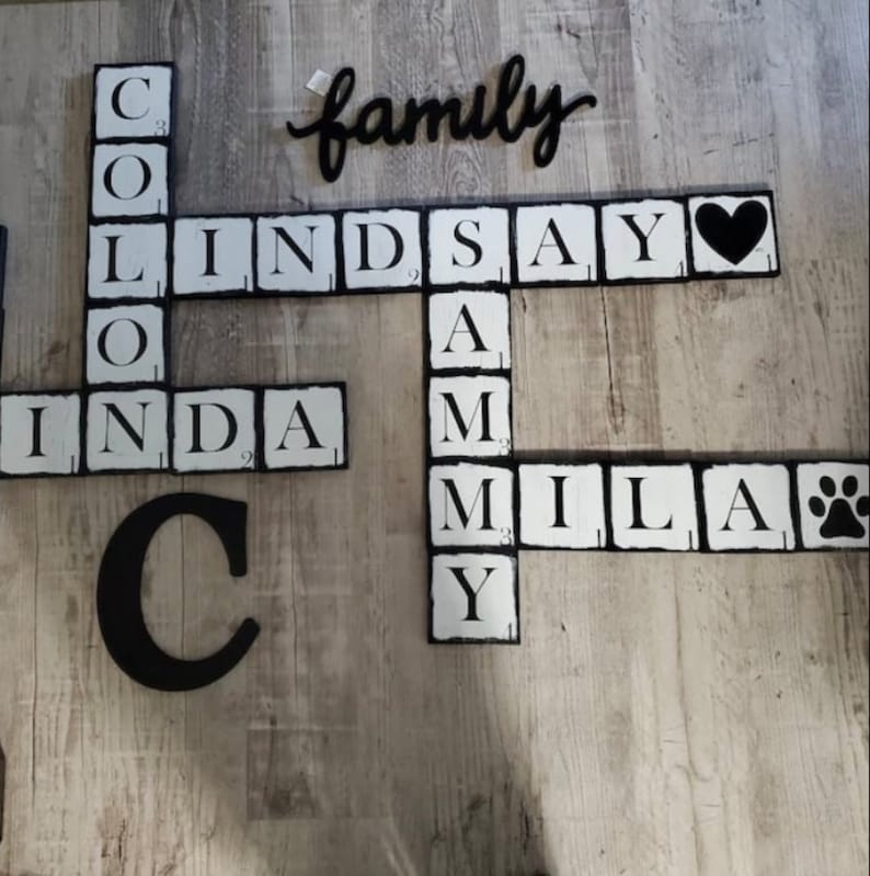 Op de afbeelding: Een houten familienaambord met zwart-witte tegels in Scrabble-stijl die namen en het woord "familie" spellen. Inclusief een zwarte hart- en pootafdruktegel. Het bord is gemonteerd op een lichte houtnerf achtergrond.