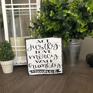 May include: A rectangular wooden sign with a distressed white finish and black edges. The sign features black hand-lettered text that reads: ACT justly LOVE mercy WALK humbly Micah 6:8. The lettering uses a mix of block and script fonts.