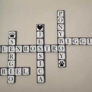 Op de afbeelding: Houten kruiswoordpuzzel muurkunst met zwarte letters op witte blokken. De puzzel bevat namen en woorden als "Ariel", "Biggie" en "Pony". Decoratieve elementen zijn een kroon, harten en pootafdrukken.