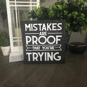以下が含まれることがあります： 「MISTAKES ARE PROOF THAT YOU'RE TRYING」と白い文字で書かれた正方形の黒い木製サイン。暗い木製の表面に置かれ、背景には白いランタン、緑、充電ステーションがあります。サインのサイズは約20cmです。