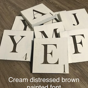 Op de afbeelding: Cr&egrave;mekleurige, verweerde houten lettertegels met donkerbruin geschilderde letters. De letters zijn gerangschikt op een houten oppervlak. De tekst "Cream distressed brown painted font" is zichtbaar.