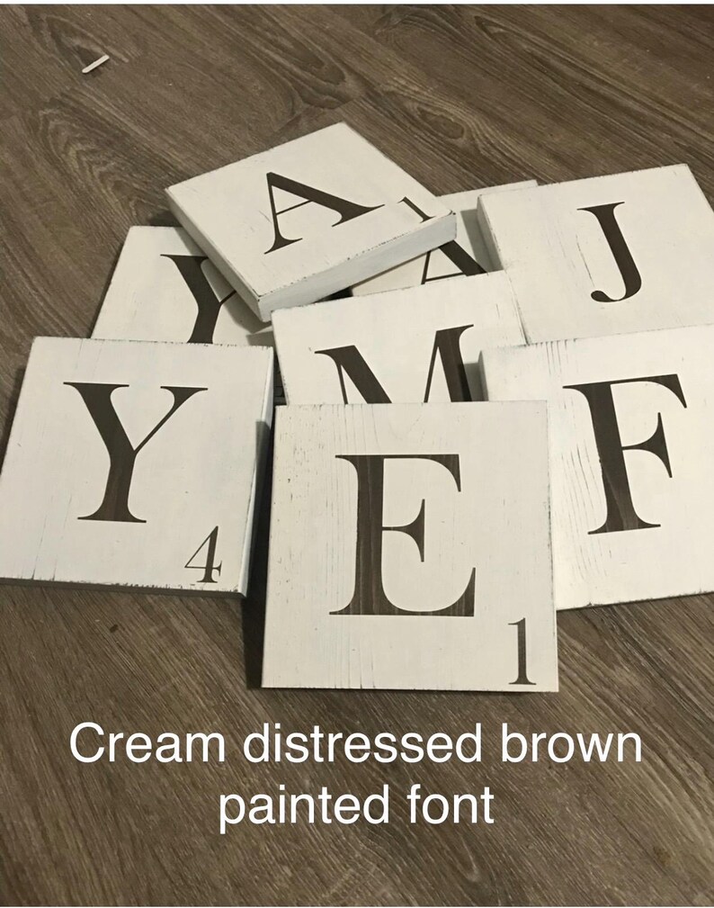 Op de afbeelding: Cr&egrave;mekleurige, verweerde houten lettertegels met donkerbruin geschilderde letters. De letters zijn gerangschikt op een houten oppervlak. De tekst "Cream distressed brown painted font" is zichtbaar.