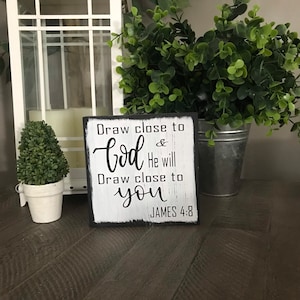 May include: A square, black-framed wooden sign with the text "Draw close to God & He will Draw close to you JAMES 4:8" in black script. The sign has a distressed, white-painted background. A white lantern and potted greenery are in the background.