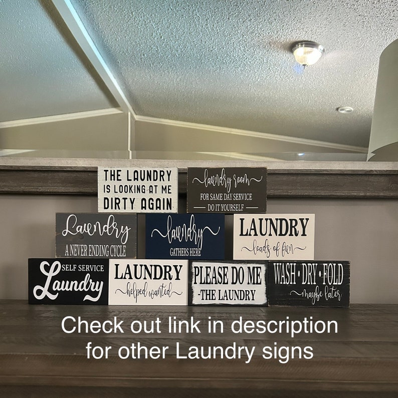 May include: A collection of small, rectangular wooden laundry signs with white lettering on various dark backgrounds. Phrases include "The Laundry is Looking at Me Dirty Again" and "Laundry Room". The bottom of the image has the text "Check out link in description for other Laundry signs."