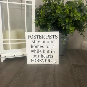 Könnte beinhalten: Ein weißes quadratisches Schild mit dem Text "FOSTER PETS stay in our homes for a while but in our hearts FOREVER" wird angezeigt. Eine Herz- und Pfotenabdruckgrafik befindet sich unten. Das Schild befindet sich auf einer Holzoberfläche, mit einer weißen Laterne und Grünzeug im Hintergrund.