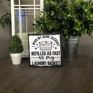 May include: A square, black-framed sign with the text "I wish my bank account refilled as fast as my laundry basket." The sign features a laundry basket graphic. The sign is on a wooden surface, with a white lantern and greenery in the background.