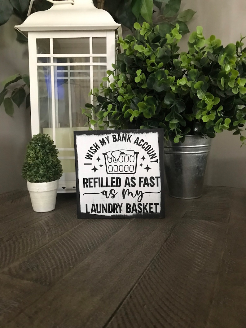 May include: A square, black-framed sign with the text "I wish my bank account refilled as fast as my laundry basket." The sign features a laundry basket graphic. The sign is on a wooden surface, with a white lantern and greenery in the background.