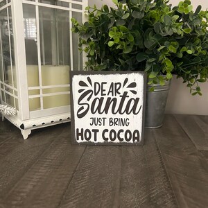 May include: A square wooden sign with a distressed white finish and black text that reads "DEAR Santa JUST BRING HOT COCOA". The sign features decorative black accents above the word "DEAR" and is positioned in front of a white lantern and a green plant.