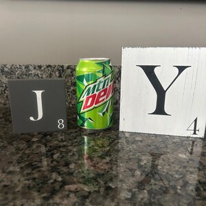 Op de afbeelding: Een groen en wit Mountain Dew frisdrankblikje staat tussen twee decoratieve lettertegels. De donkergrijze tegel links heeft een witte "J" en het nummer "8". De witte tegel rechts heeft een zwarte "Y" en het nummer "4".