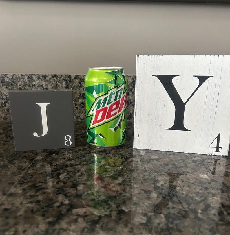 Op de afbeelding: Een groen en wit Mountain Dew frisdrankblikje staat tussen twee decoratieve lettertegels. De donkergrijze tegel links heeft een witte "J" en het nummer "8". De witte tegel rechts heeft een zwarte "Y" en het nummer "4".