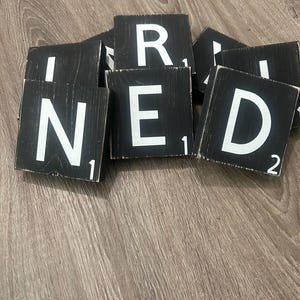 May include: Black wooden Scrabble tiles with white letters spelling "I NEED". Each tile has a number in the corner. The tiles are arranged on a wooden surface.