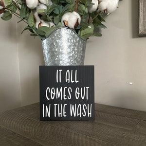 May include: A black wooden sign with white text that reads "IT ALL COMES OUT IN THE WASH" sits on a wooden surface. Behind the sign is a galvanized metal bucket filled with cotton stems and green leaves.