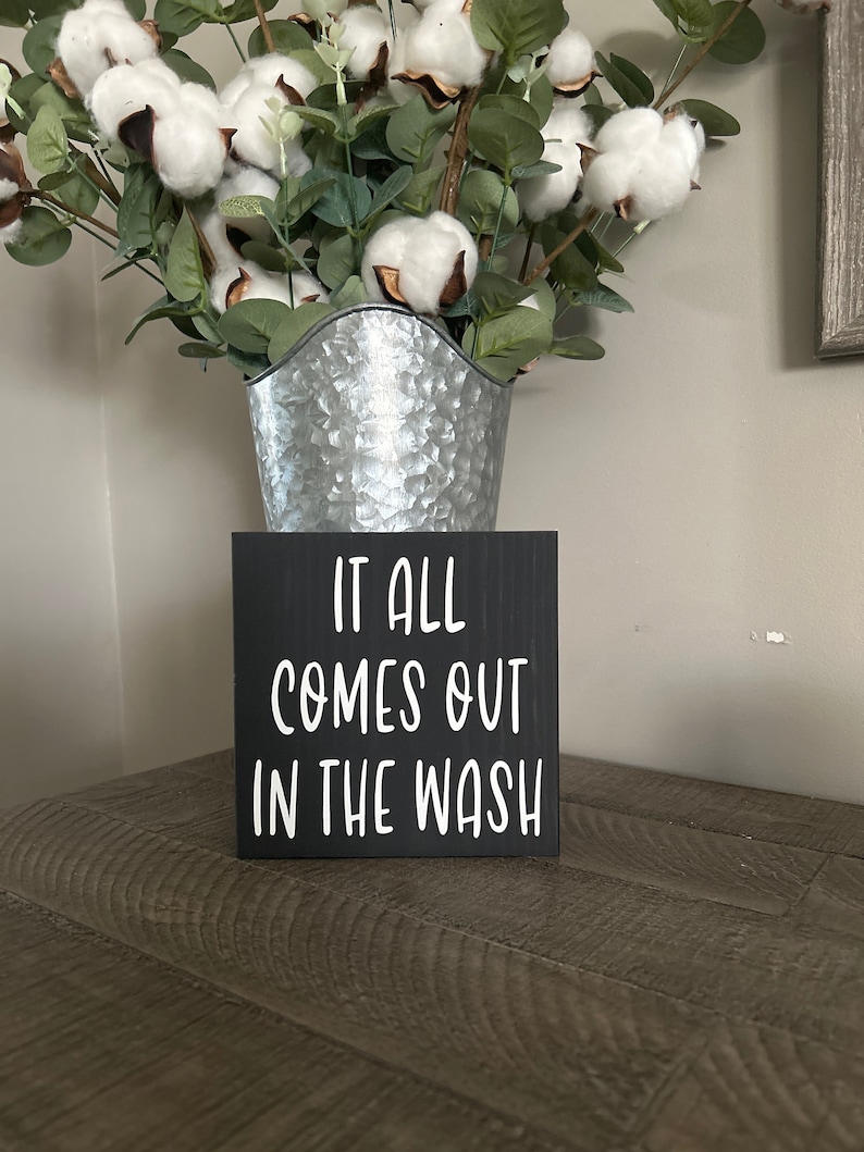 May include: A black wooden sign with white text that reads "IT ALL COMES OUT IN THE WASH" sits on a wooden surface. Behind the sign is a galvanized metal bucket filled with cotton stems and green leaves.
