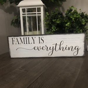 Könnte beinhalten: Ein weißes Holzschild mit schwarzem Text, der "FAMILY IS everything" lautet. Das Schild steht auf einer dunklen Holzoberfläche mit einer weißen Laterne und grünen Pflanzen im Hintergrund.