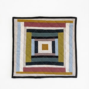 Könnte beinhalten: Quadratischer, gesteppter Wandbehang mit geometrischem Design. Der Quilt hat ein zentrales Quadrat in Gold- und Brauntönen, umgeben von konzentrischen Rechtecken in verschiedenen Farben, darunter Blau, Grün und Burgunderrot. Der Quilt ist schwarz eingefasst.