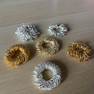 May include: Six metallic finger rings in silver and gold tones are arranged on a light wood surface. The rings have a coiled design, with a central opening. The rings are likely used for stress relief or as a fashion accessory.