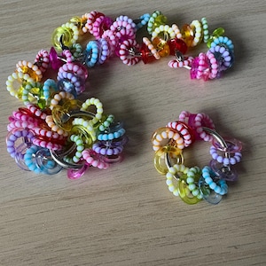 May include: A selection of colourful beaded rings and a bracelet. The beads are in various colours, including pink, yellow, blue, and green. The rings and bracelet are connected by silver-coloured metal rings. The items are arranged on a light brown wooden surface.
