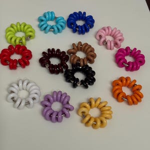 May include: A collection of thirteen circular hair ties in various colours including lime green, light blue, dark blue, pink, red, dark brown, tan, magenta, white, purple, black, orange, and yellow, each featuring a coiled plastic design.