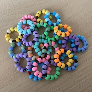 Può includere: Una collezione di anelli di perline colorate, ognuno realizzato con piccole perline rotonde in varie tonalità di blu, verde, giallo, arancione, rosa e viola. Gli anelli sono impilati uno sopra l'altro, creando una visualizzazione vibrante e giocosa.