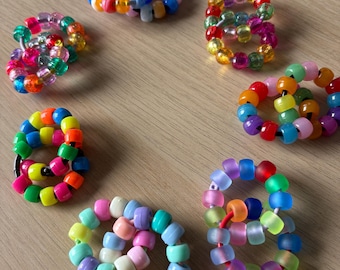 Può includere: Una collezione di anelli di perline colorate in varie dimensioni e combinazioni di colori. Gli anelli presentano una varietà di colori, tra cui rosso, arancione, giallo, verde, blu e viola. Le perline sono disposte in un motivo circolare.