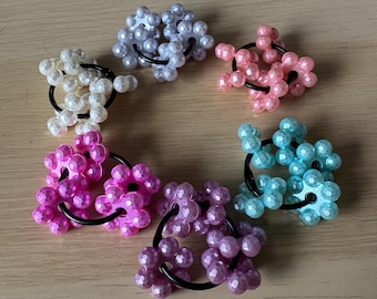 Può includere: Sei elastici per capelli con perline in vari colori, tra cui bianco, rosa, viola e blu. Ogni elastico presenta un gruppo di piccole perline rotonde attaccate a un elastico nero. Le perline hanno una finitura lucida e perlescente.