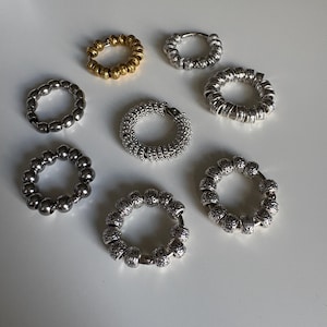 May include: A collection of seven circular hoop earrings arranged on a surface. The earrings feature various metallic finishes including gold and silver, with some designs showcasing smooth spherical beads and others displaying intricate engraved patterns.