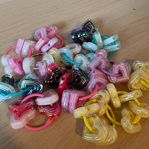 Può includere: Una collezione di elastici colorati per capelli con perline a forma di cuore. Gli elastici presentano una varietà di colori, tra cui rosa, giallo, blu e iridescente. Le perline sono realizzate in un materiale traslucido e sono disposte su elastici.